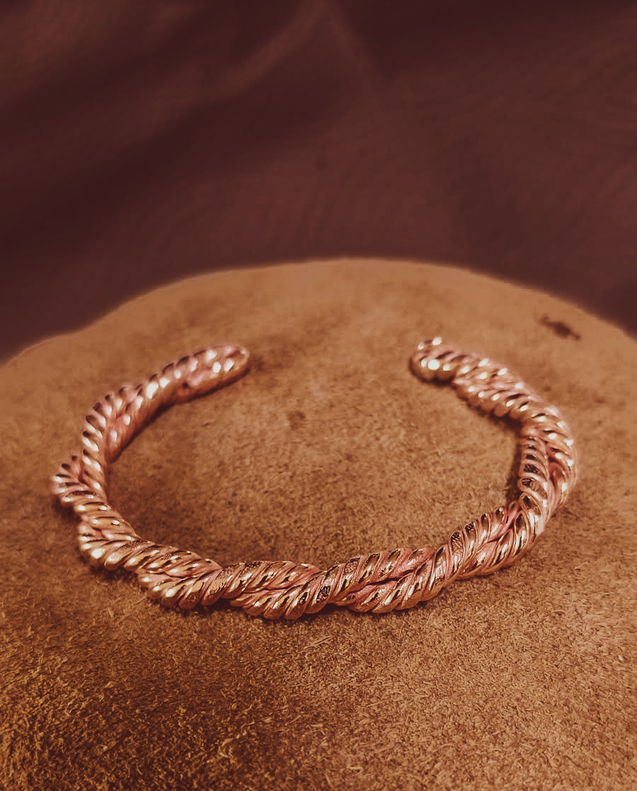 A twisted rope-style cuff bracelet hand forged in copper, displayed on a textured brown surface with a dark brown background.