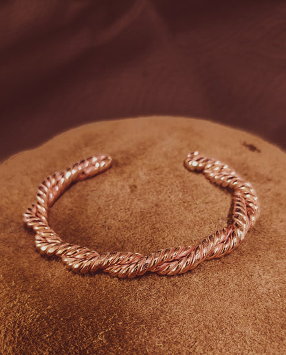 A twisted rope-style cuff bracelet hand forged in copper, displayed on a textured brown surface with a dark brown background.