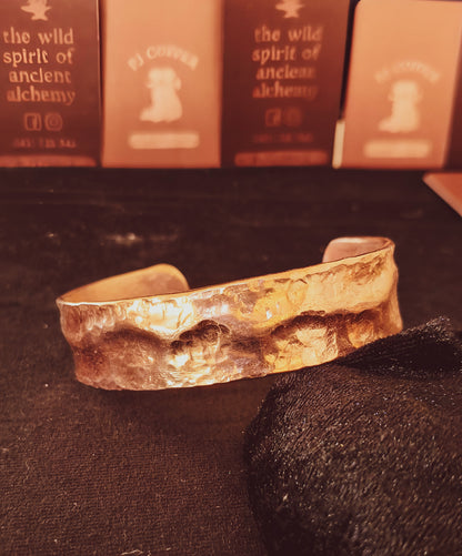 A hammered copper cuff bracelet, resting on a dark textured surface with a black cloth in the foreground and branded cards blurred in the background.