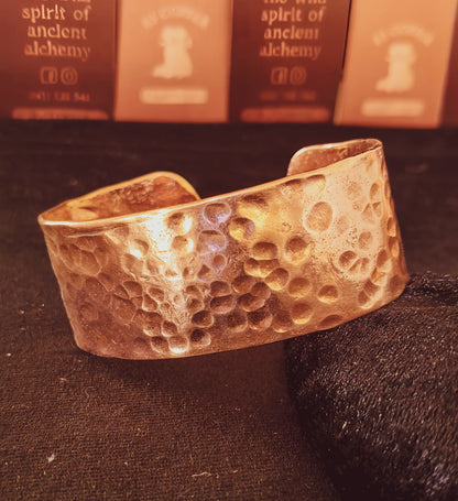 A hammered copper cuff bracelet with moon-like craters, resting on a dark textured surface with blurred books in the background.