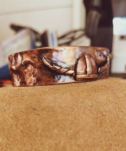 A hammered copper cuff bracelet with a central viking motif, resting on a textured tan surface with a blurred indoor background.