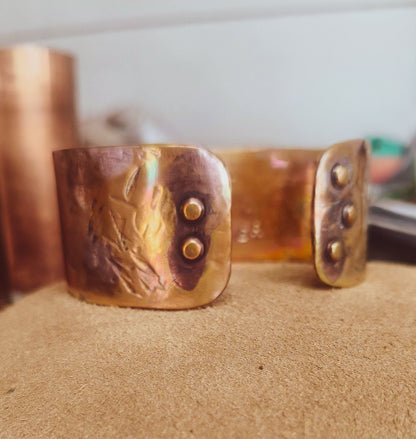 A hammered copper cuff bracelet with brass rivets, resting on a textured tan surface with a blurred background.