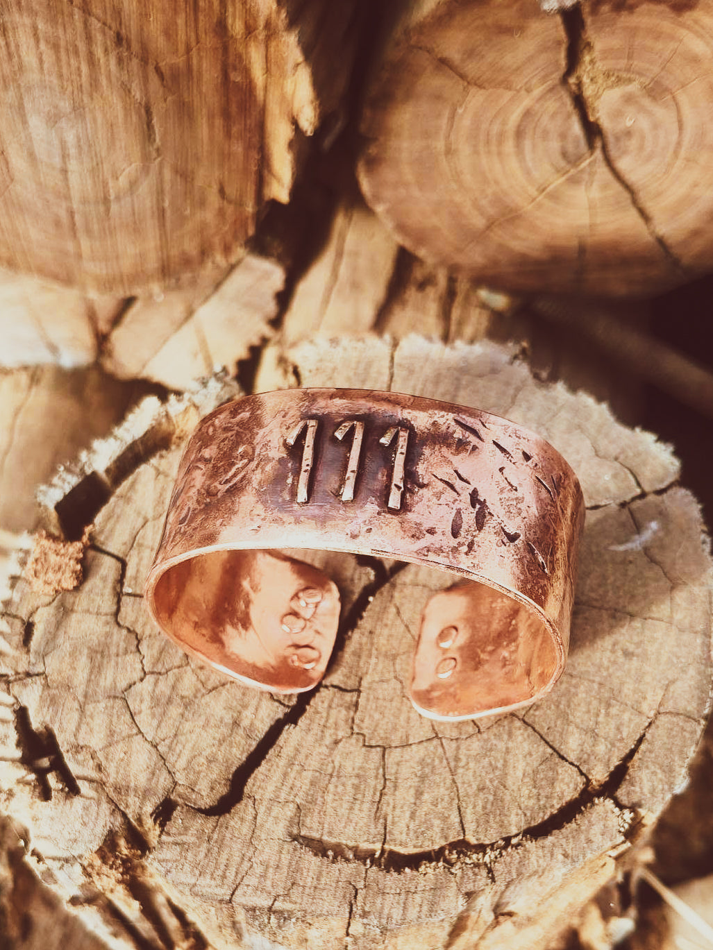 A rustic copper cuff bracelet with stamped details and the number '111' on the front, displayed on a weathered wood log.
