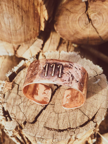 A rustic copper cuff bracelet with stamped details and the number '111' on the front, displayed on a weathered wood log.