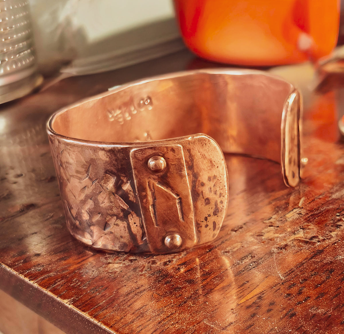 A hammered copper cuff bracelet with a decorative front tab secured by two rivets, resting on a rustic wooden tabletop.