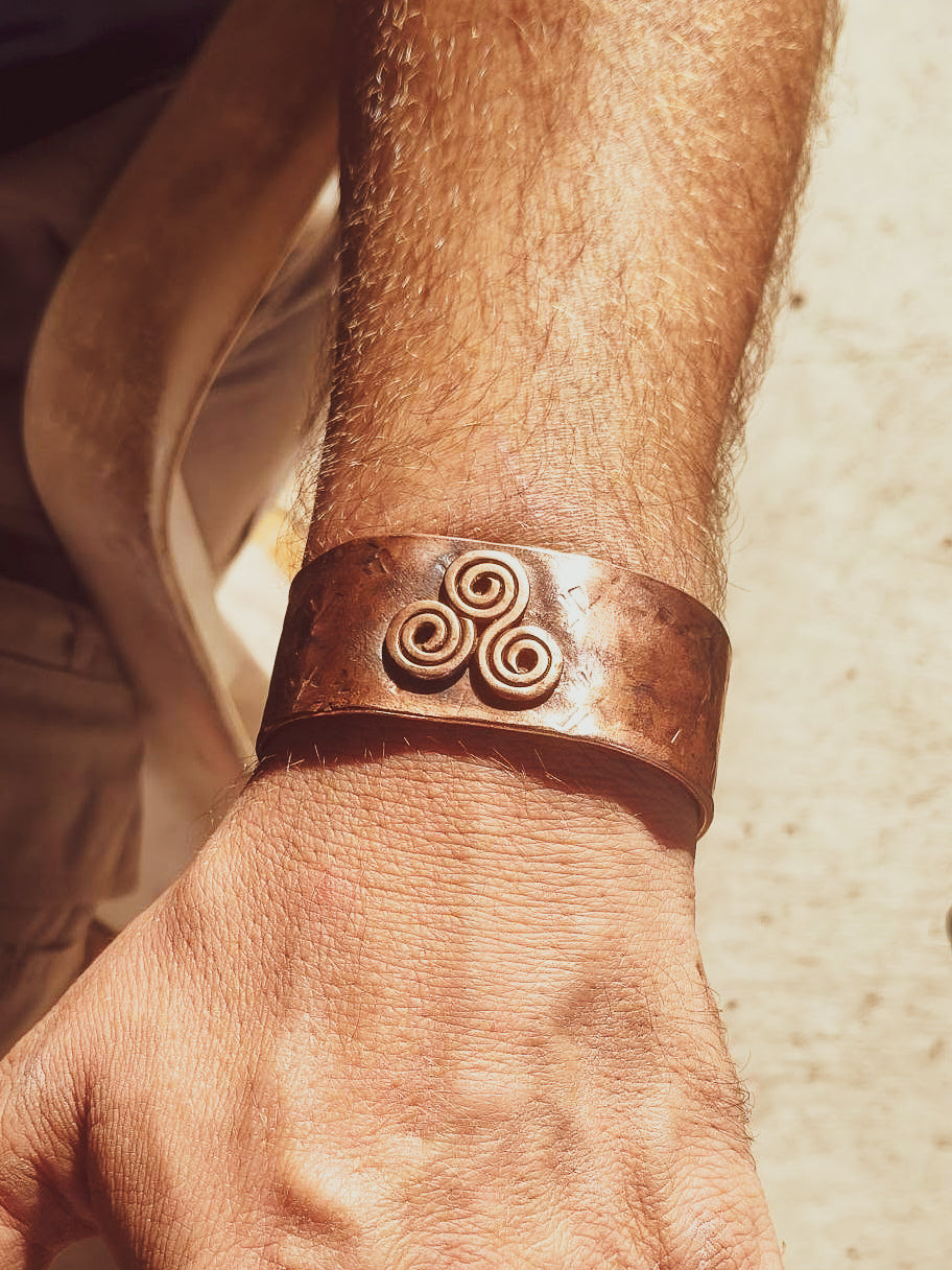 A close-up of a person's wrist wearing a wide, hammered copper cuff bracelet with a raised triskelion motif, photographed in warm natural light.