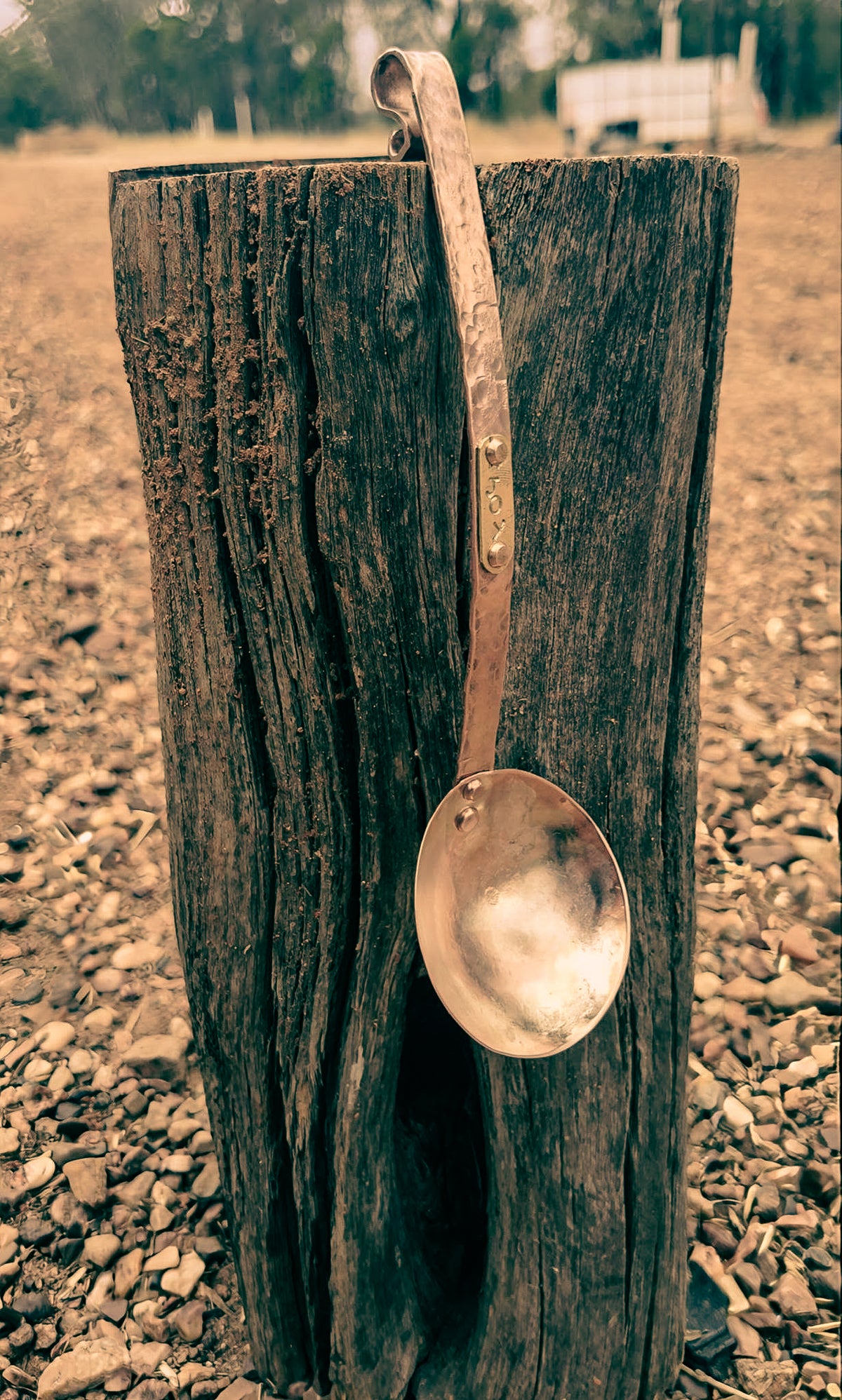 A rustic copper ladle with a hammered handle and spoon bowl, hanging from a weathered wooden post outdoors on gravel ground.