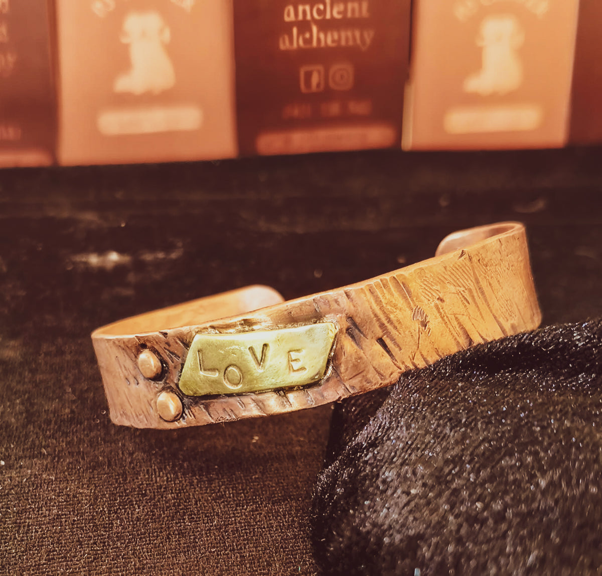 A handmade tree bark textured copper cuff bracelet with a stamped metal plate reading 'LOVE' and two small rivet details, resting on a dark textured surface with blurred cards in the background.