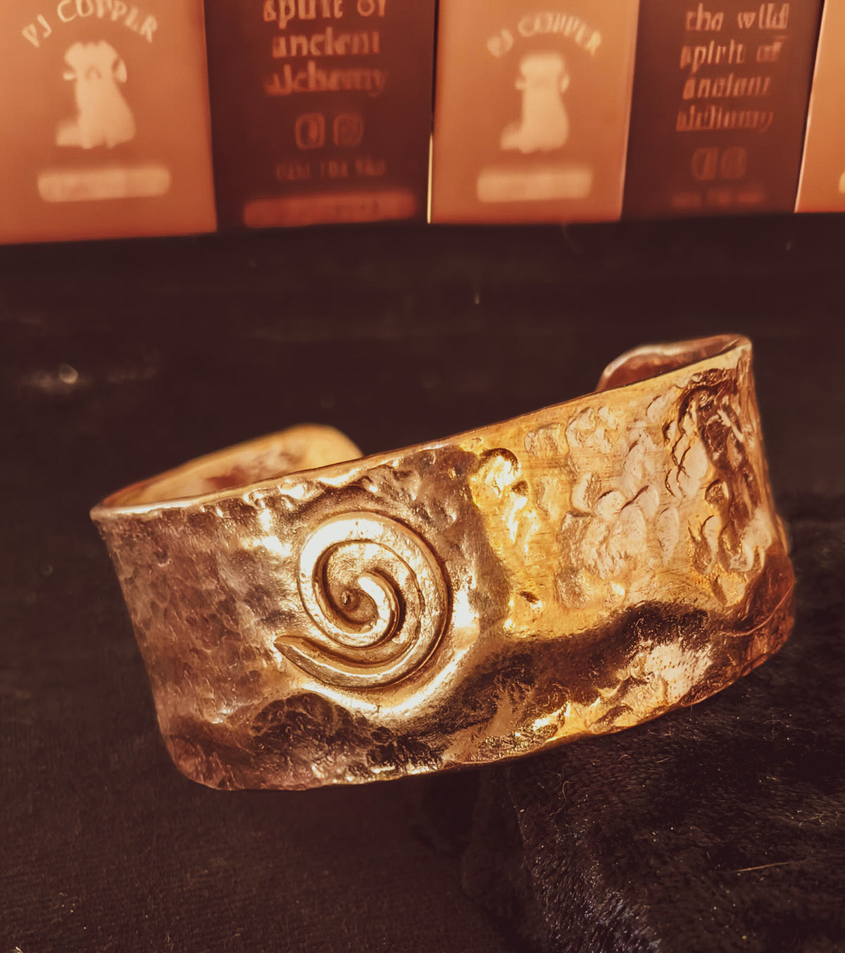 A hammered copper cuff bracelet with a spiral motif, photographed on a dark surface with warm amber-toned cards in the background.