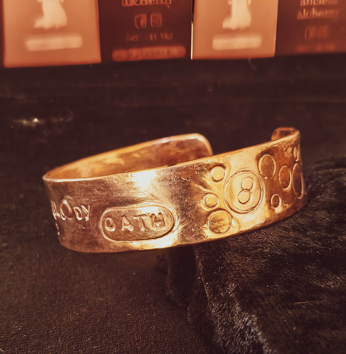 A textured copper cuff bracelet with engraved names and numbers, resting on a black velvet surface with warm-toned cards blurred in the background.