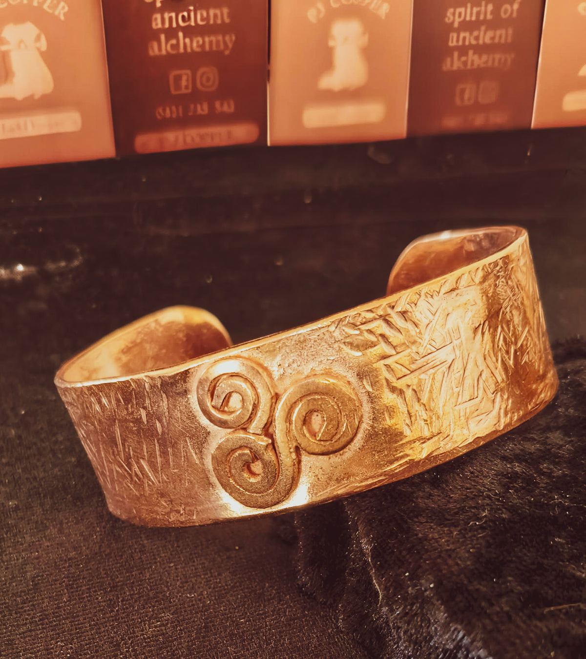 A textured copper metail cuff bracelet with a Triskelion symbol, resting on a dark fabric surface with PJ Copper cards in background