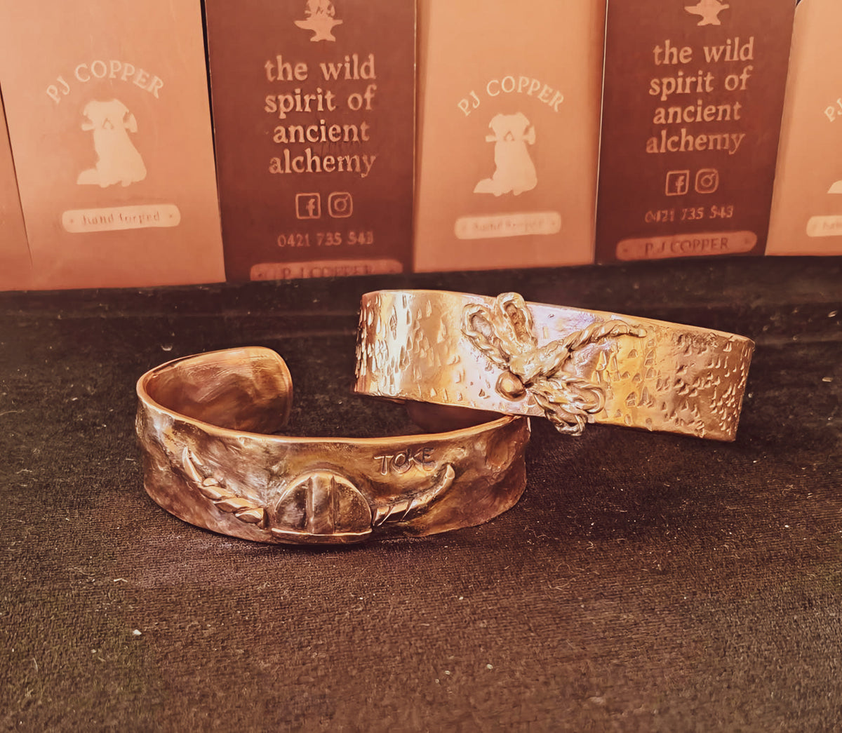 Two hammered metal cuff bracelets in warm copper displayed on a dark surface, with branded cards in the background reading 'PJ COPPER' and 'the wild spirit of ancient alchemy'.