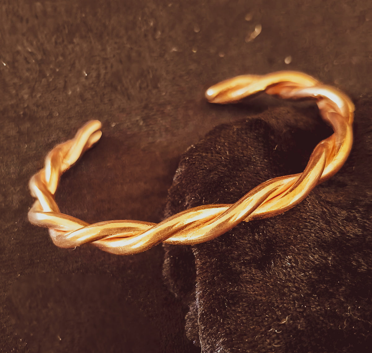 A twisted open cuff bracelet in warm copper photographed close-up against a dark textured background with a black fabric accent.