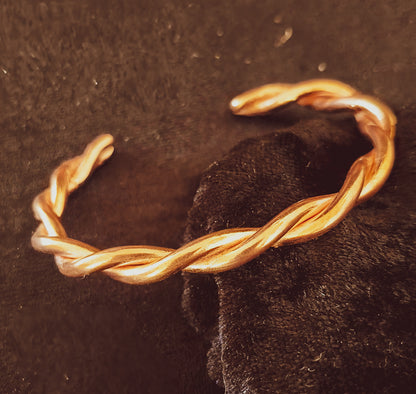 A twisted open cuff bracelet in warm copper photographed close-up against a dark textured background with a black fabric accent.